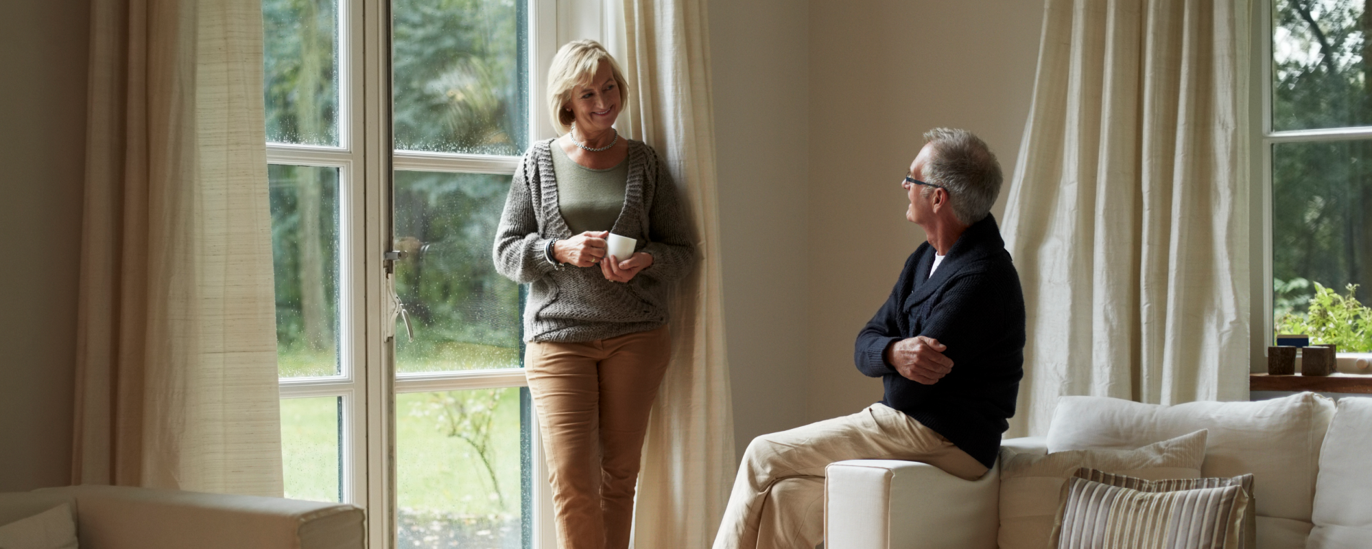 Man and a woman holding a coffee cup in their living room smiling at each other by the window