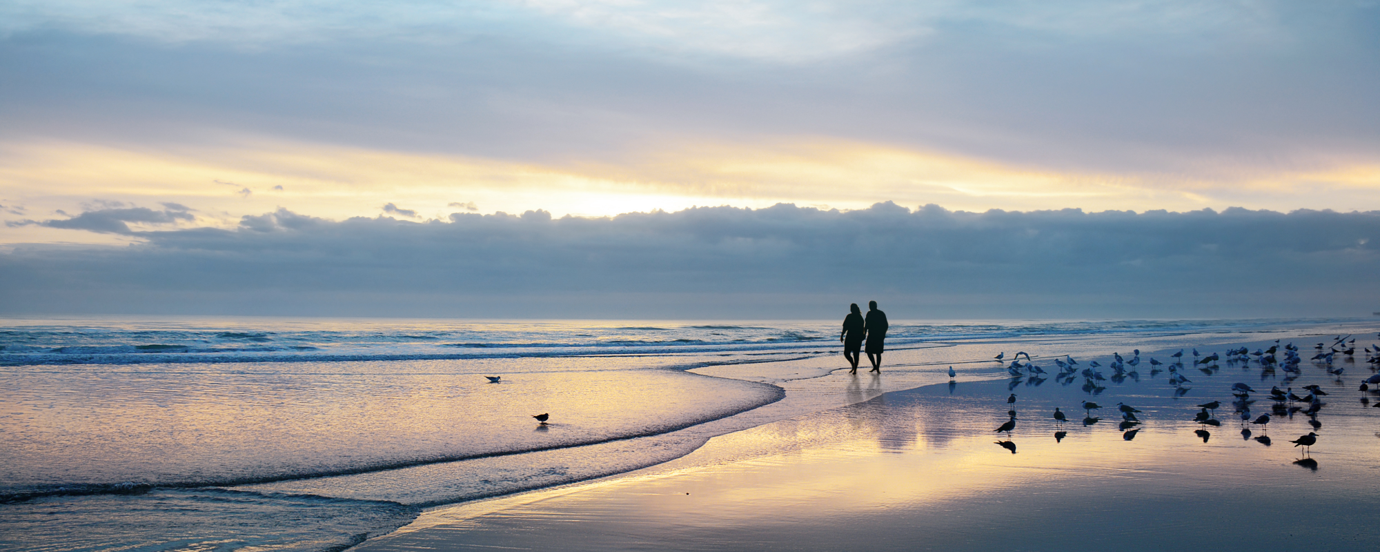 Elderly couple walk along a beach shore at sunset