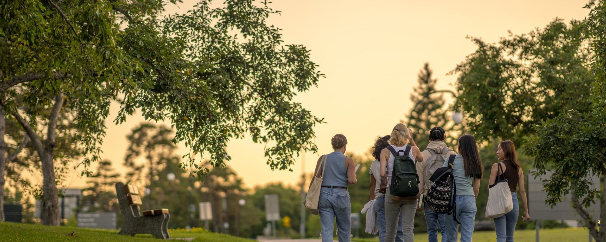 A group of University students are seen from behind walking outside on campus as they make their way to class. They are dressed casually and have backpacks on as they talk together and make their way to school for the day..