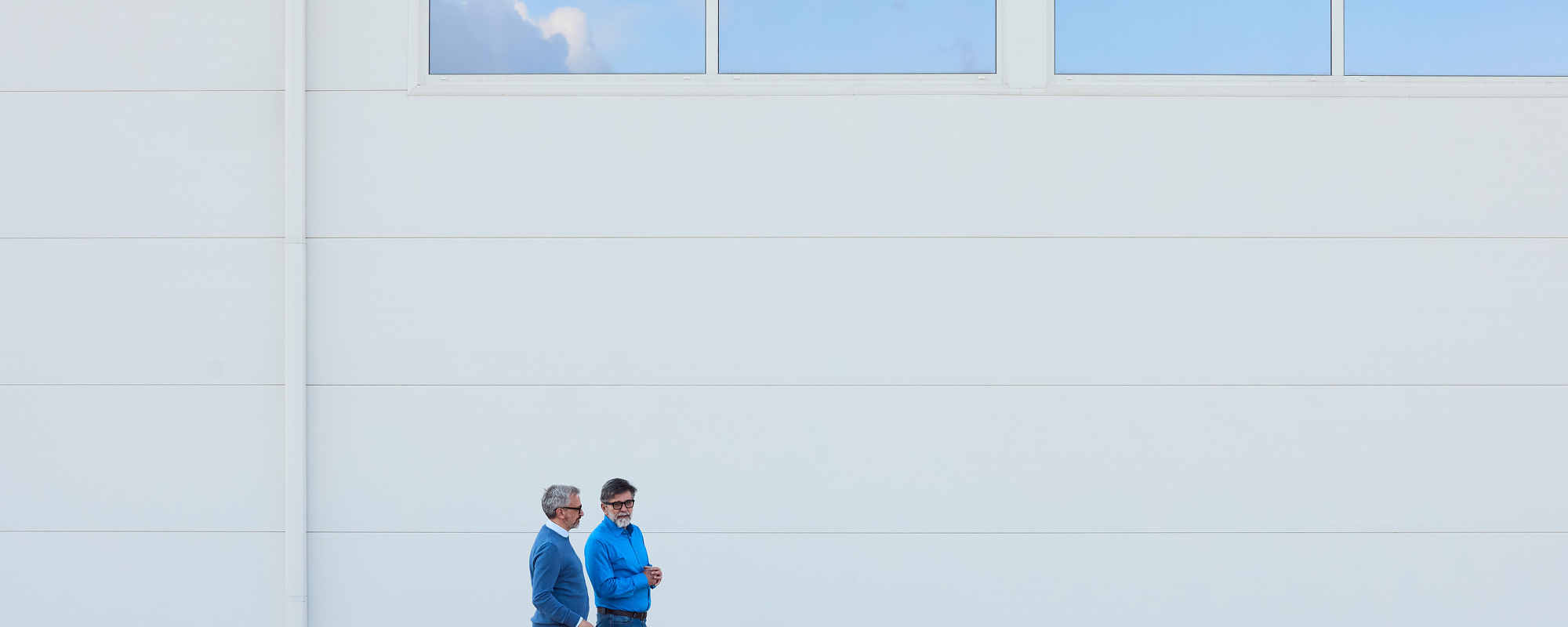 Two individuals walk near a modern white building with clear architectural lines..